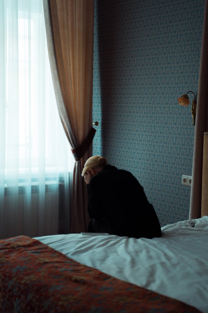 gallery-1 A solitary woman sits on a bed, gazing out a window, evoking feelings of solitude and introspection.